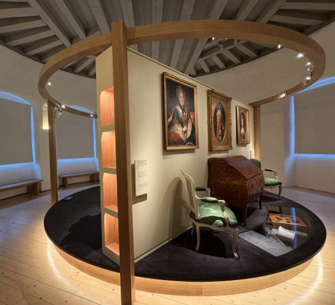 Salle d’exposition muséale présentant des portraits anciens, un bureau d’époque et deux fauteuils.